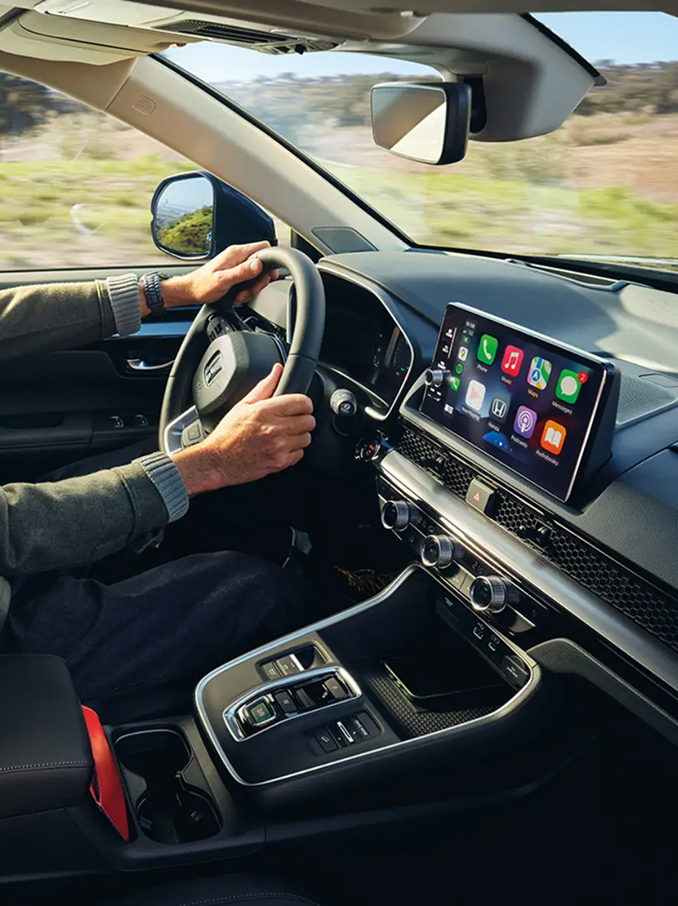 Vue de l'habitacle d'un véhicule moderne avec un conducteur installé derrière le volant, montrant le tableau de bord, le volant, la console centrale et un large écran tactile affichant des icônes d'applications colorées, tandis qu'un paysage de campagne défile par les fenêtres.