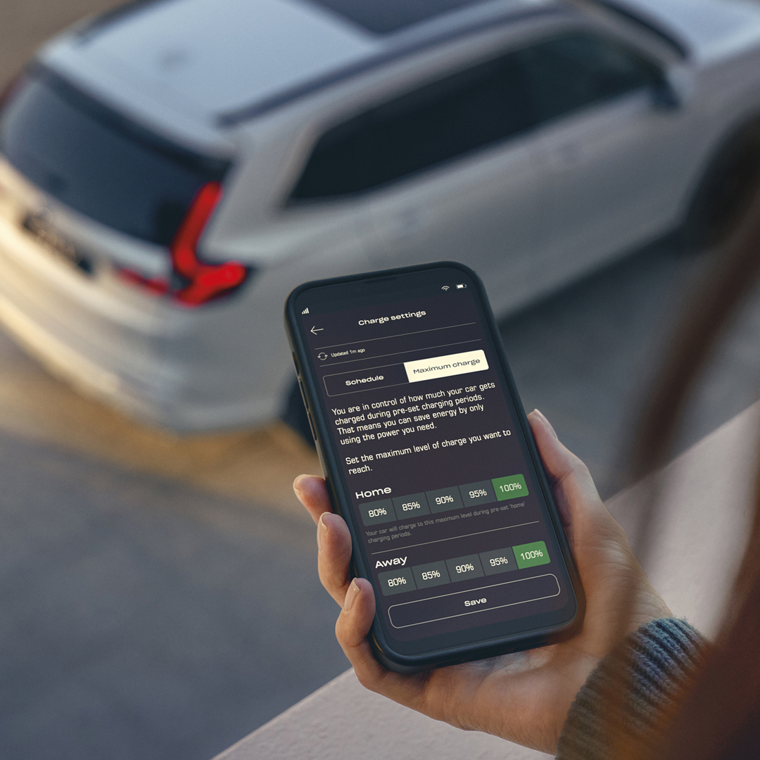 Une personne tient un smartphone affichant une application de gestion de la recharge du véhicule, tandis qu'un SUV blanc est garé en arrière-plan.