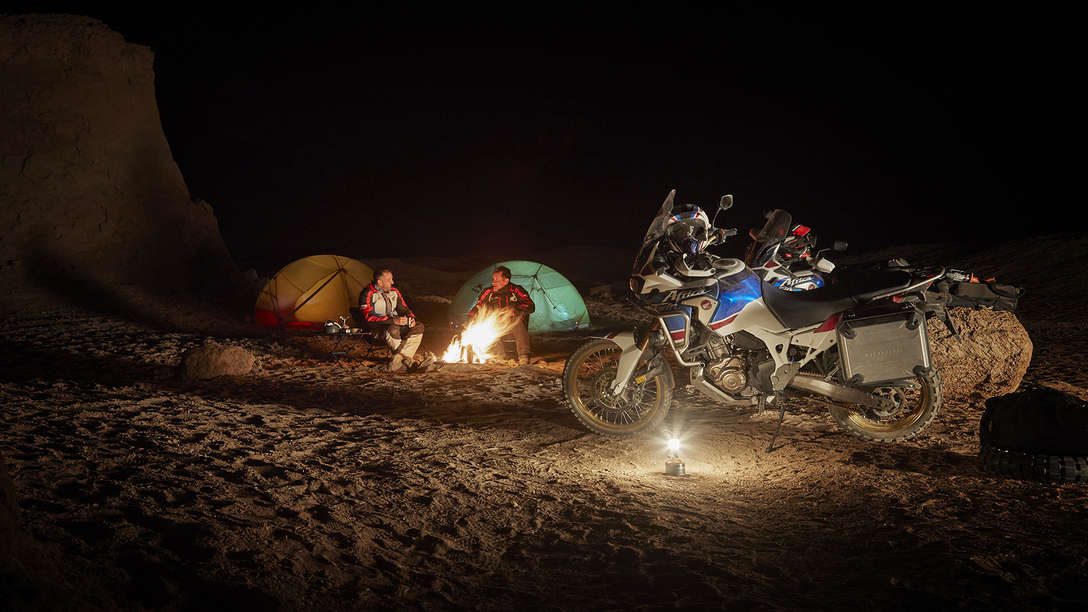 Deux personnes faisant du camping et vue lat&eacute;rale du trail Honda Africa Twin Adventure Sports stationn&eacute;e, de nuit.