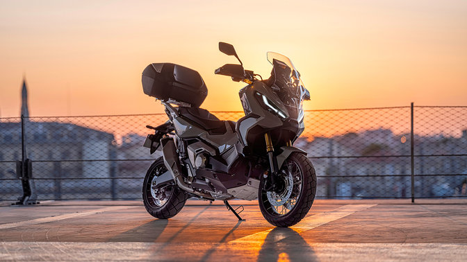 Vue de trois quarts avant droit de la Honda X-ADV, grise, à l'arrêt en stationnement, avec un coucher de soleil
