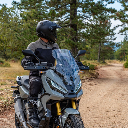 Vue de trois quarts avant droit de la Honda X-ADV, grise, avec pilote debout sur la moto, sur un chemin forestier