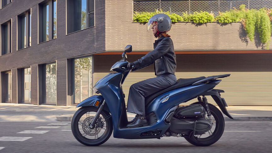Femme au guidon d’un Honda Sh350i, de profil, en milieu urbain.