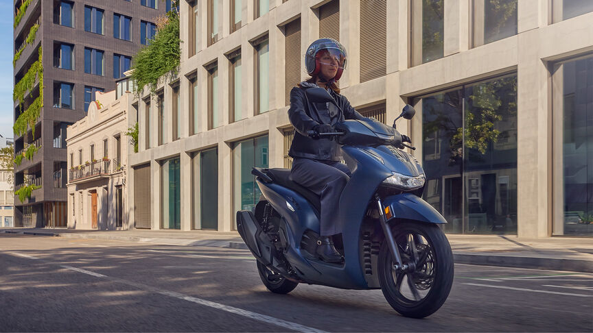 Femme au guidon d’un Honda Sh350i, de profil, en milieu urbain.